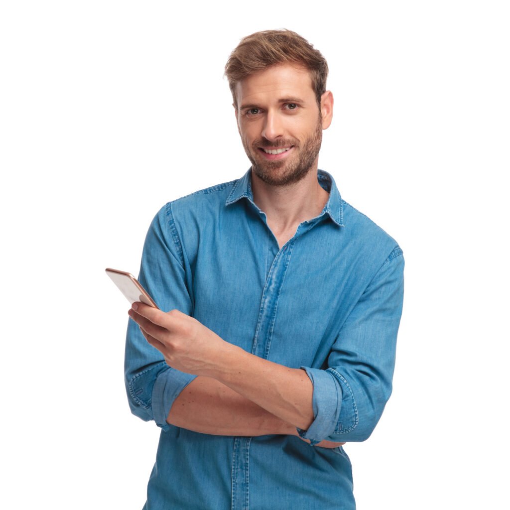 Smiling man in a blue denim shirt holds a smartphone while crossing his arms.