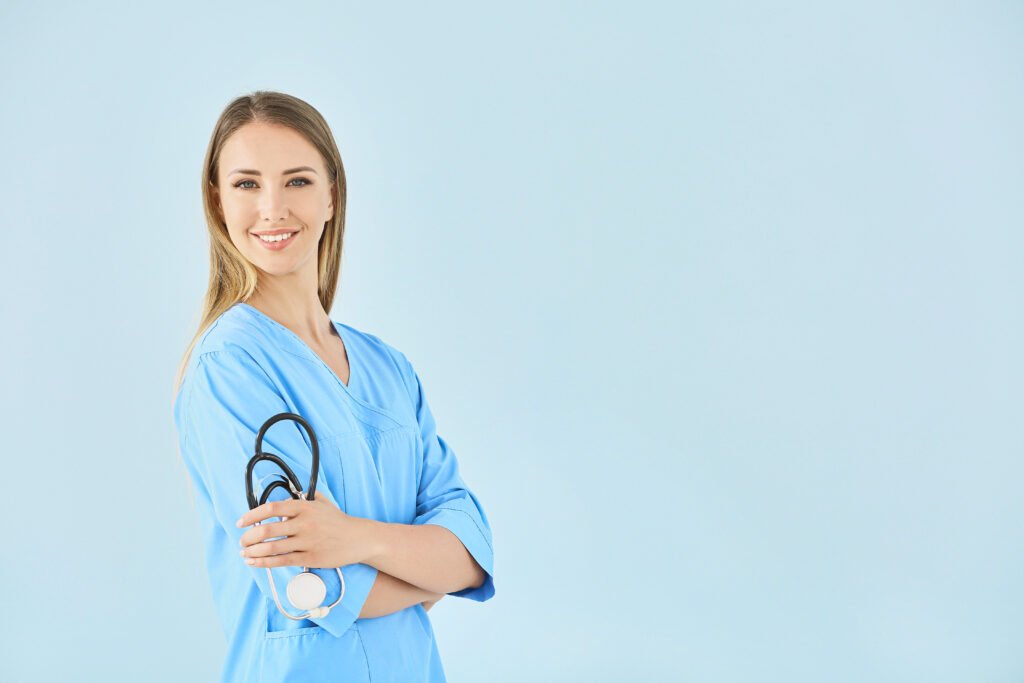 Female nurse in blue scrubs with stethoscope, arms crossed, smiling against a light blue background.